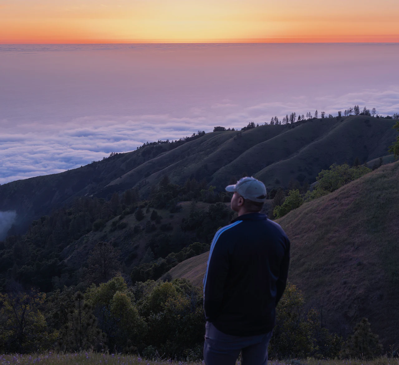 A man in a ball cap stands on a hillside, looking out over rolling foothills and a sea of clouds at sunset.