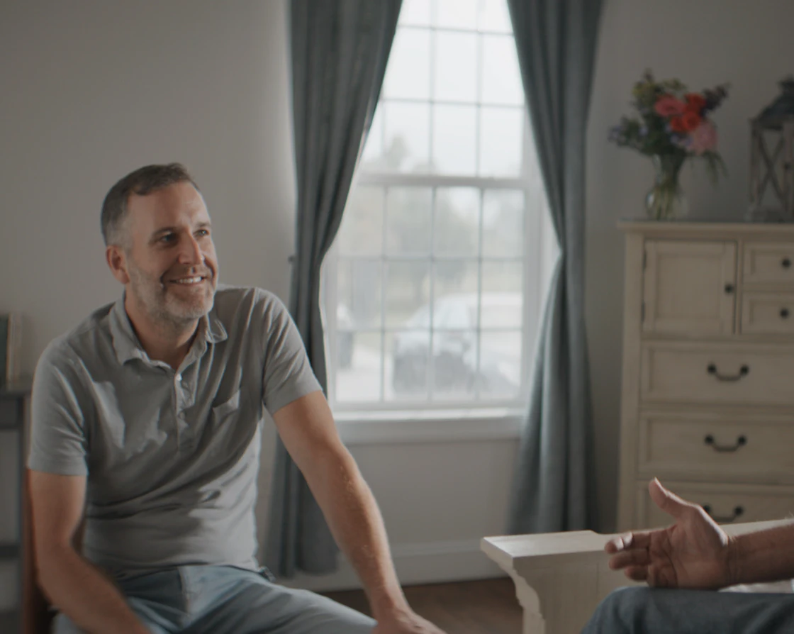 A smiling man talks with an older man in a calm, sunlit room.